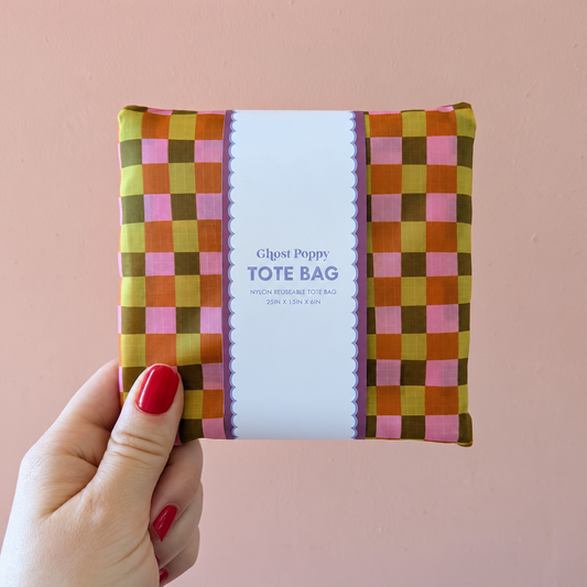 A hand holds a folded Fall Plaid Reusable Tote Bag in an orange, olive, and pink plaid pattern, wrapped in a paper sleeve labeled with the product name and dimensions.