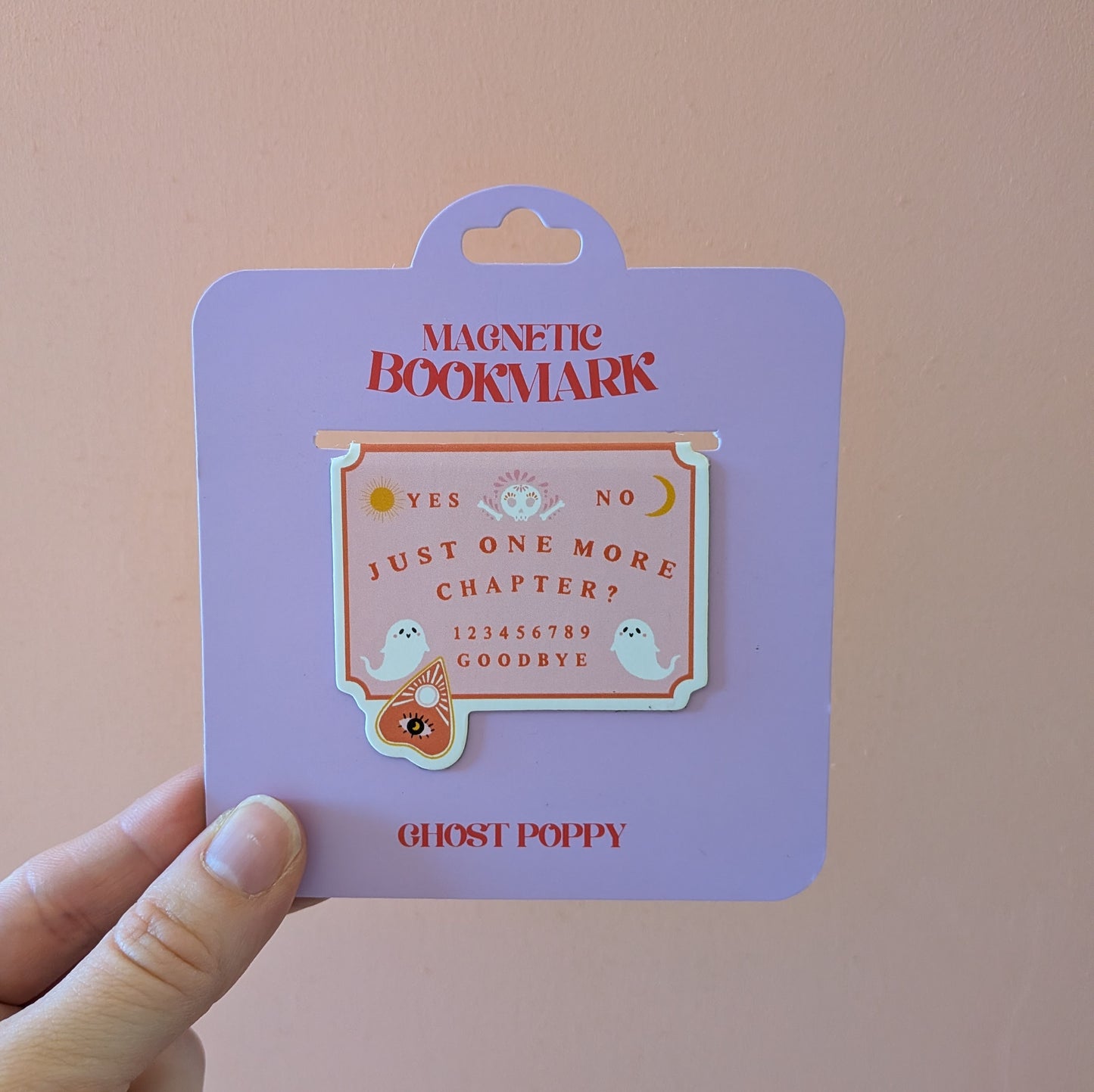 A hand holds a purple card featuring the “Just One More Chapter? Magnetic Bookmark” by Ghost Poppy—a whimsical, Ouija board-shaped page marker adorned with small ghost illustrations and the brand name “Ghost Poppy” at the bottom. Perfect for any book lover seeking unique magnetic bookmarks.