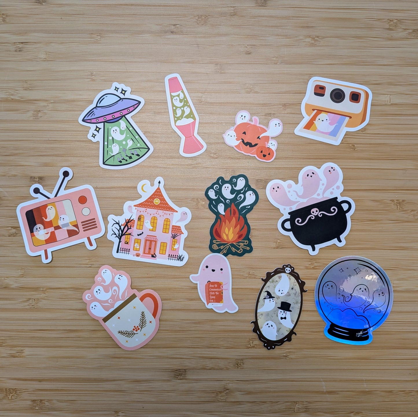 Bright, assorted vinyl stickers from the Ghost Poppy “Mystery Ghost Sticker Pack” showcase adorable ghosts in fun settings—like a UFO, lava lamp, campfire, cauldron, and spooky house—all artistically displayed on a wooden surface. Perfect for decorating your favorite items or adding to your own mystery sticker collection!.