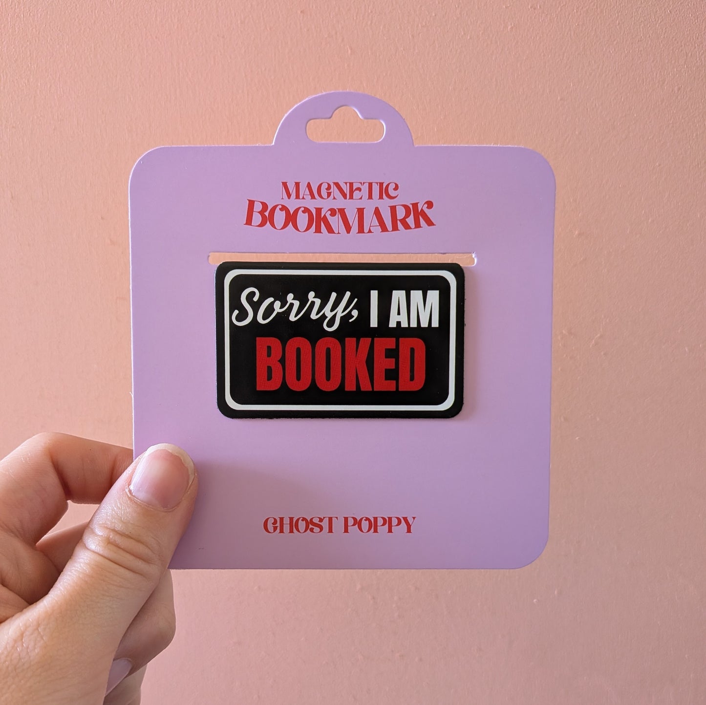 A hand holds the Ghost Poppy Sorry I am Booked Magnetic Bookmark, displayed on lavender packaging that features the phrase “Sorry, I am BOOKED” in black, white, and red text. The brand name appears at the bottom, making this giftable bookmark both practical and stylish.