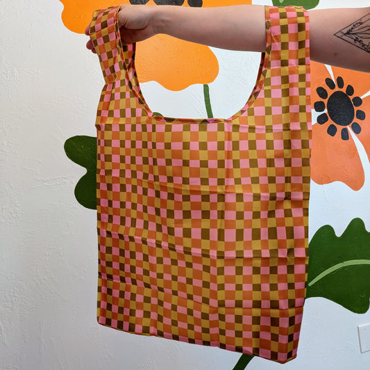 A hand holds up a Ghost Poppy Fall Plaid Reusable Tote Bag featuring a pink, yellow, and brown checkered pattern.