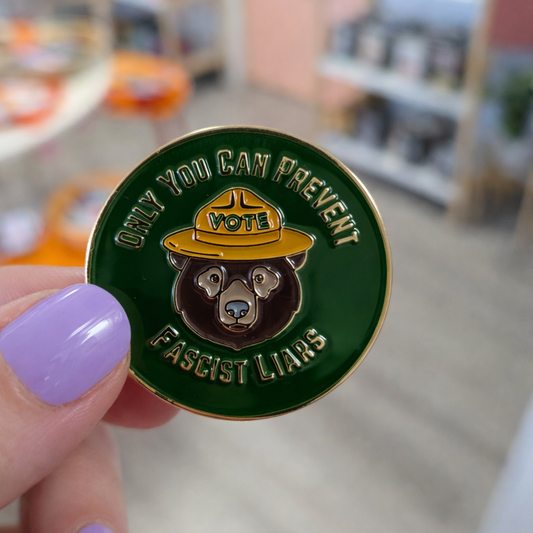 Pin with bear design and text held by a hand in a blurred indoor setting