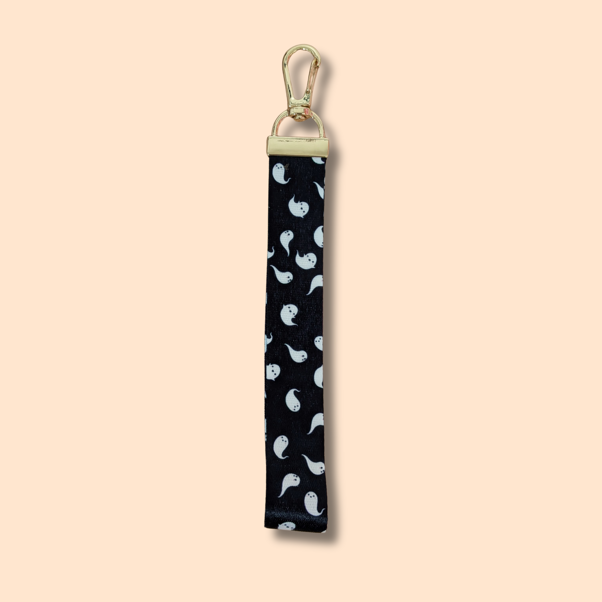 The Ghost Parade Wristlet Lanyard Keychain by Ghost Poppy features a black fabric strap adorned with a pattern of small white ghosts and is finished with a gold clasp, shown against a beige background.