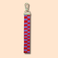 The Ghost Poppy Red & Purple Wristlet Lanyard Keychain, featuring a gold lobster clasp and a red and purple checkered fabric strap, hangs vertically against a light peach background.