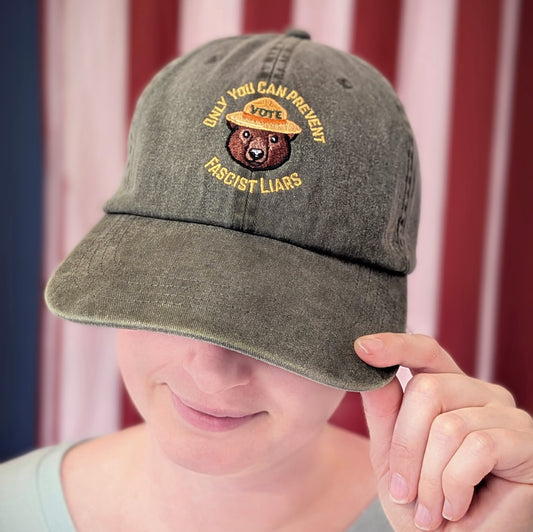 A person wears a grey Only You Dad Hat by Ghost Poppy, embroidered with a bear in a park ranger hat that says Only You Can Prevent Fascist Liars and Vote.