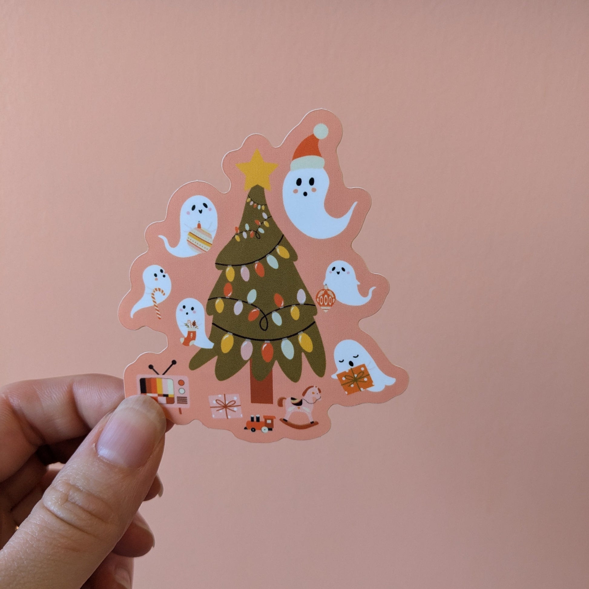 A hand holds the Ghost Poppy “Yuletide Hauntings Sticker,” a matte vinyl Christmas sticker showcasing a Christmas tree adorned with lights, surrounded by cute cartoon ghosts, presents, and small festive items, set against a peach-colored background.