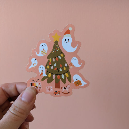 A hand holds the Ghost Poppy “Yuletide Hauntings Sticker,” a matte vinyl Christmas sticker showcasing a Christmas tree adorned with lights, surrounded by cute cartoon ghosts, presents, and small festive items, set against a peach-colored background.
