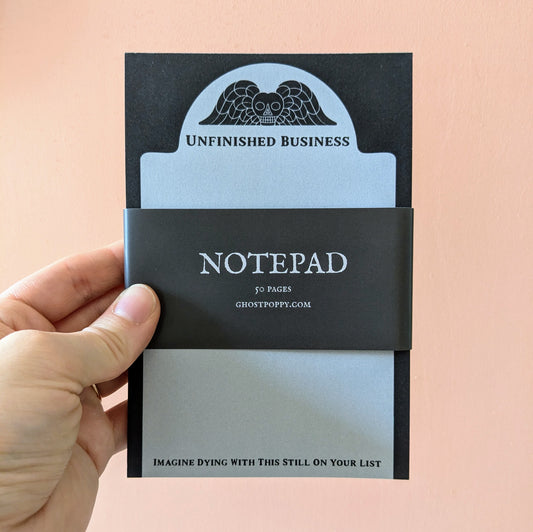 A hand holds the Ghost Poppy Unfinished Business Notepad, a 4x6″ pad featuring an owl and wings illustration. This distinctive stationery offers 50 pages and showcases the bold phrase: IMAGINE DYING WITH THIS STILL ON YOUR LIST.