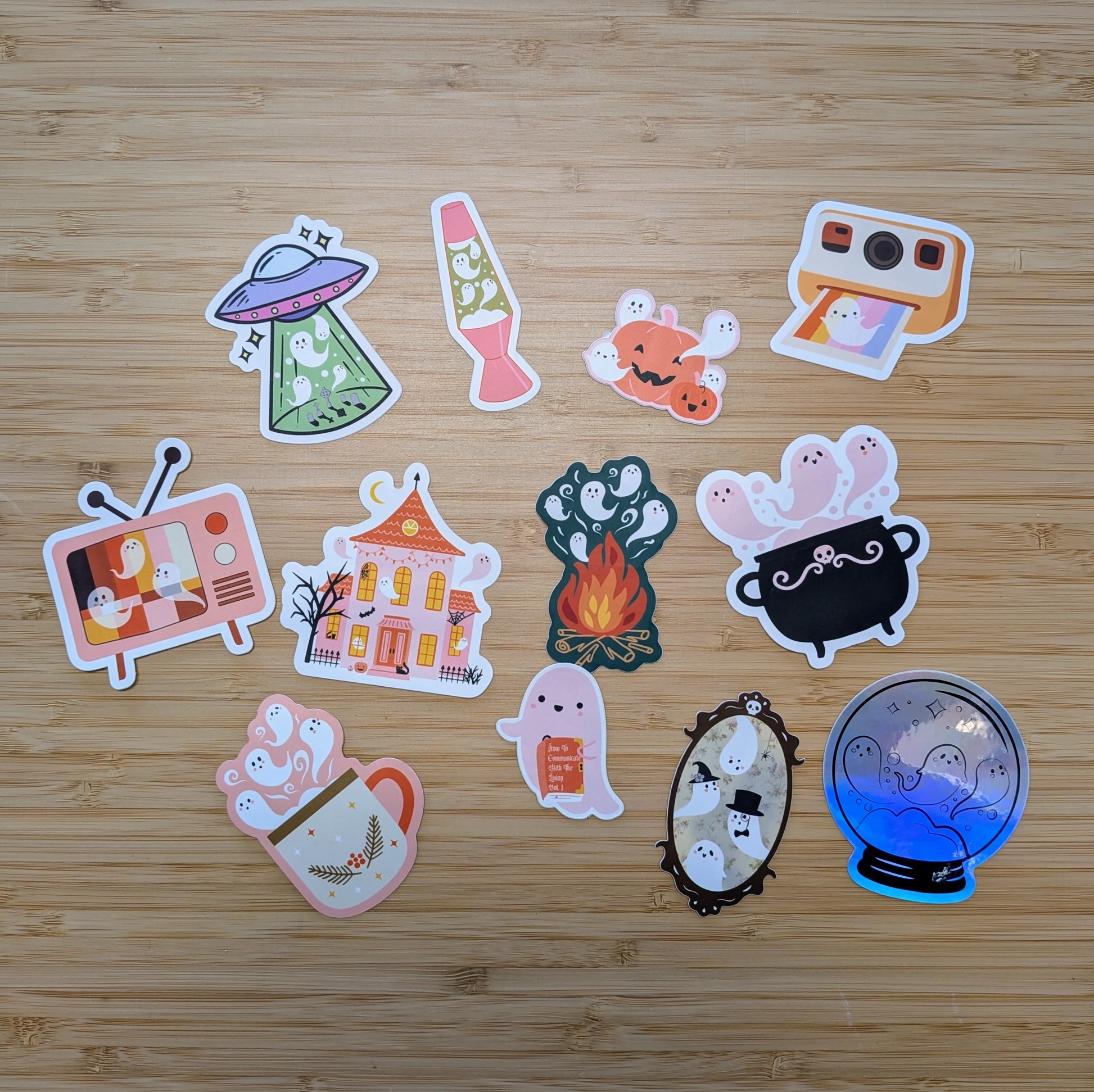 Bright, assorted vinyl stickers from the Ghost Poppy “Mystery Ghost Sticker Pack” showcase adorable ghosts in fun settings—like a UFO, lava lamp, campfire, cauldron, and spooky house—all artistically displayed on a wooden surface. Perfect for decorating your favorite items or adding to your own mystery sticker collection!.