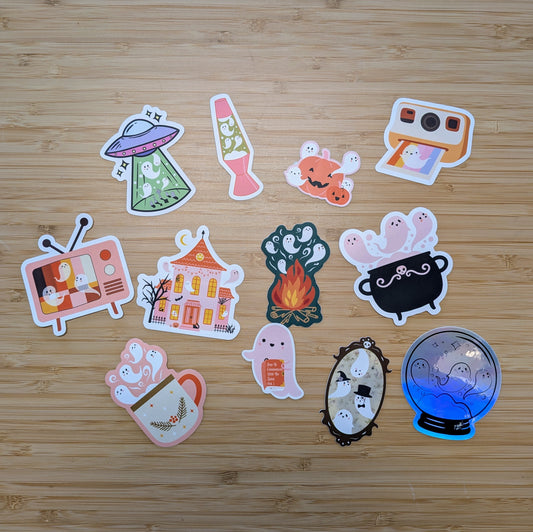 Bright, assorted vinyl stickers from the Ghost Poppy “Mystery Ghost Sticker Pack” showcase adorable ghosts in fun settings—like a UFO, lava lamp, campfire, cauldron, and spooky house—all artistically displayed on a wooden surface. Perfect for decorating your favorite items or adding to your own mystery sticker collection!.