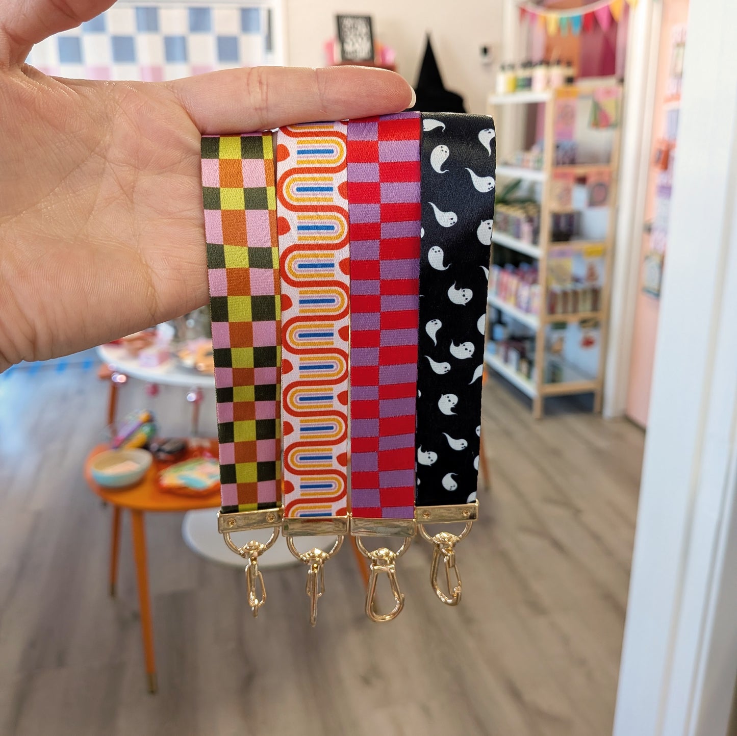 A hand displays four Ghost Poppy Rainbow Wristlet Lanyard Keychains in assorted patterns—pink and green checker, orange and yellow squiggles, red checker, and black with white ghosts—all finished with gold lobster clasps. A lively boutique setting appears in the background.