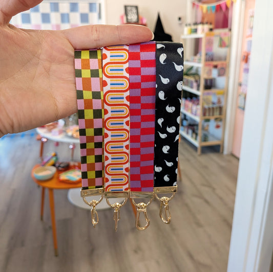 A hand holds four colorful Fall Plaid Wristlet Lanyard Keychains from Ghost Poppy, each featuring a unique pattern: pink-green checkered, retro orange with numun, red checkered, and black with white ghost shapes. In the background is a playful, colorful room.