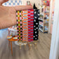 A hand holds four colorful Fall Plaid Wristlet Lanyard Keychains from Ghost Poppy, each featuring a unique pattern: pink-green checkered, retro orange with numun, red checkered, and black with white ghost shapes. In the background is a playful, colorful room.