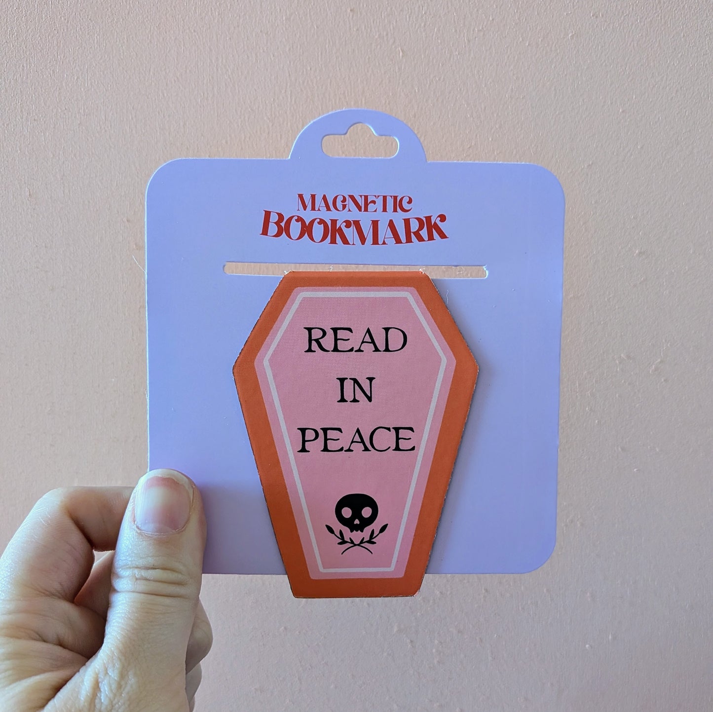 A hand holds the Ghost Poppy Read In Peace Magnetic Bookmark—an eye-catching, coffin-shaped accessory featuring a skull and the phrase “READ IN PEACE.” The packaging displays “MAGNETIC BOOKMARK” at the top.