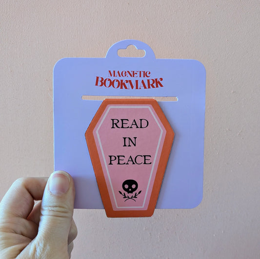 A hand holds the Ghost Poppy Read In Peace Magnetic Bookmark—an eye-catching, coffin-shaped accessory featuring a skull and the phrase “READ IN PEACE.” The packaging displays “MAGNETIC BOOKMARK” at the top.