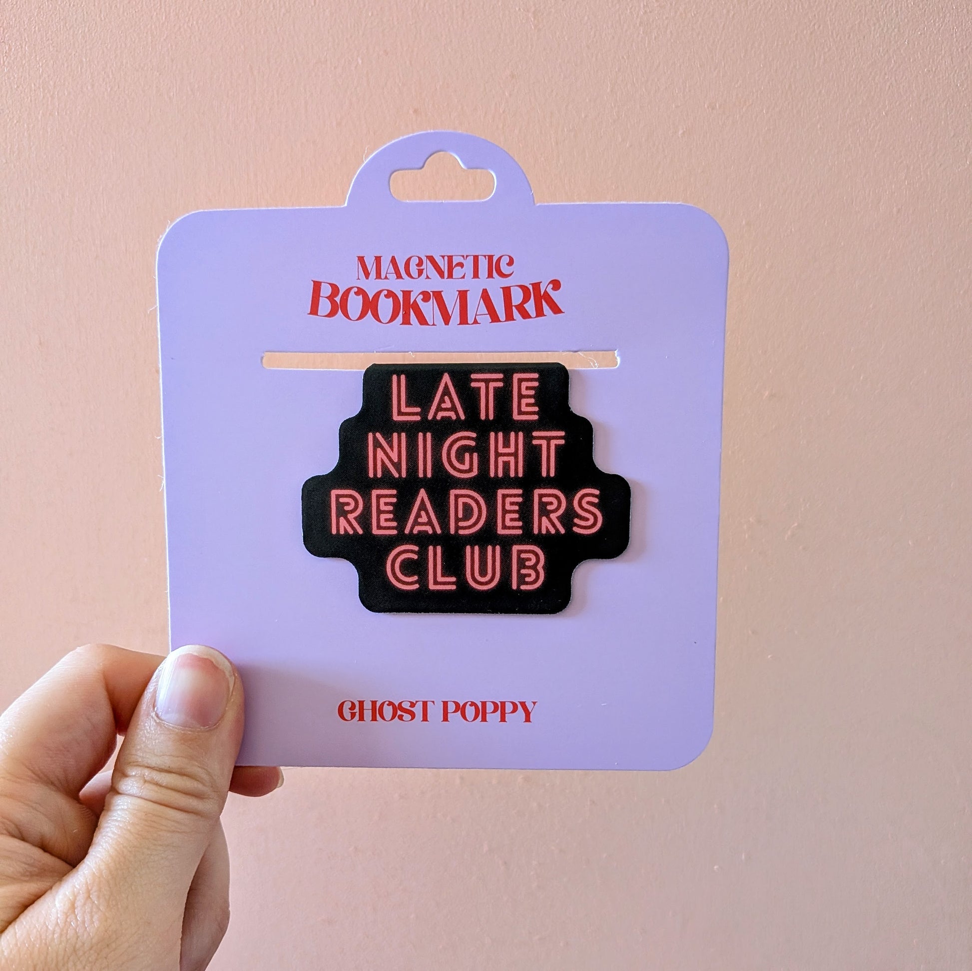 A hand holds the Late Night Readers Club Magnetic Bookmark by Ghost Poppy, displayed on lavender card packaging. This stylish black magnetic bookmark, featuring “LATE NIGHT READERS CLUB” in neon pink letters, is a perfect accessory for books or planners. The card reads “MAGNETIC BOOKMARK” and “GHOST POPPY.”.