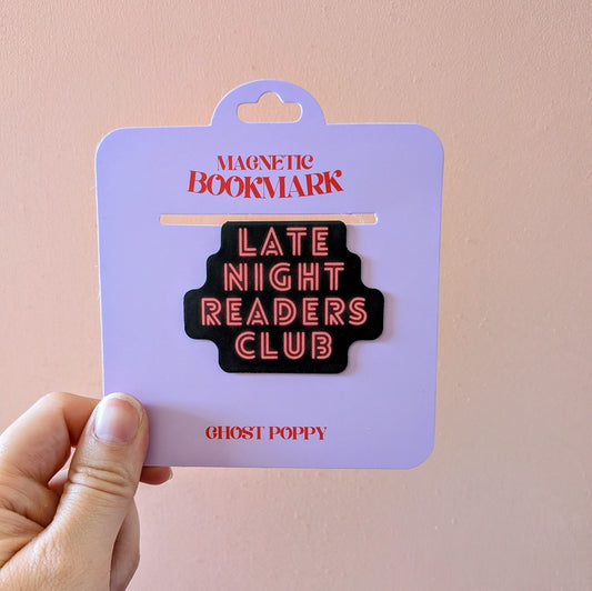 A hand holds the Late Night Readers Club Magnetic Bookmark by Ghost Poppy, displayed on lavender card packaging. This stylish black magnetic bookmark, featuring “LATE NIGHT READERS CLUB” in neon pink letters, is a perfect accessory for books or planners. The card reads “MAGNETIC BOOKMARK” and “GHOST POPPY.”.