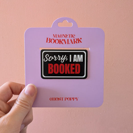 A hand holds the Ghost Poppy Sorry I am Booked Magnetic Bookmark, displayed on lavender packaging that features the phrase “Sorry, I am BOOKED” in black, white, and red text. The brand name appears at the bottom, making this giftable bookmark both practical and stylish.