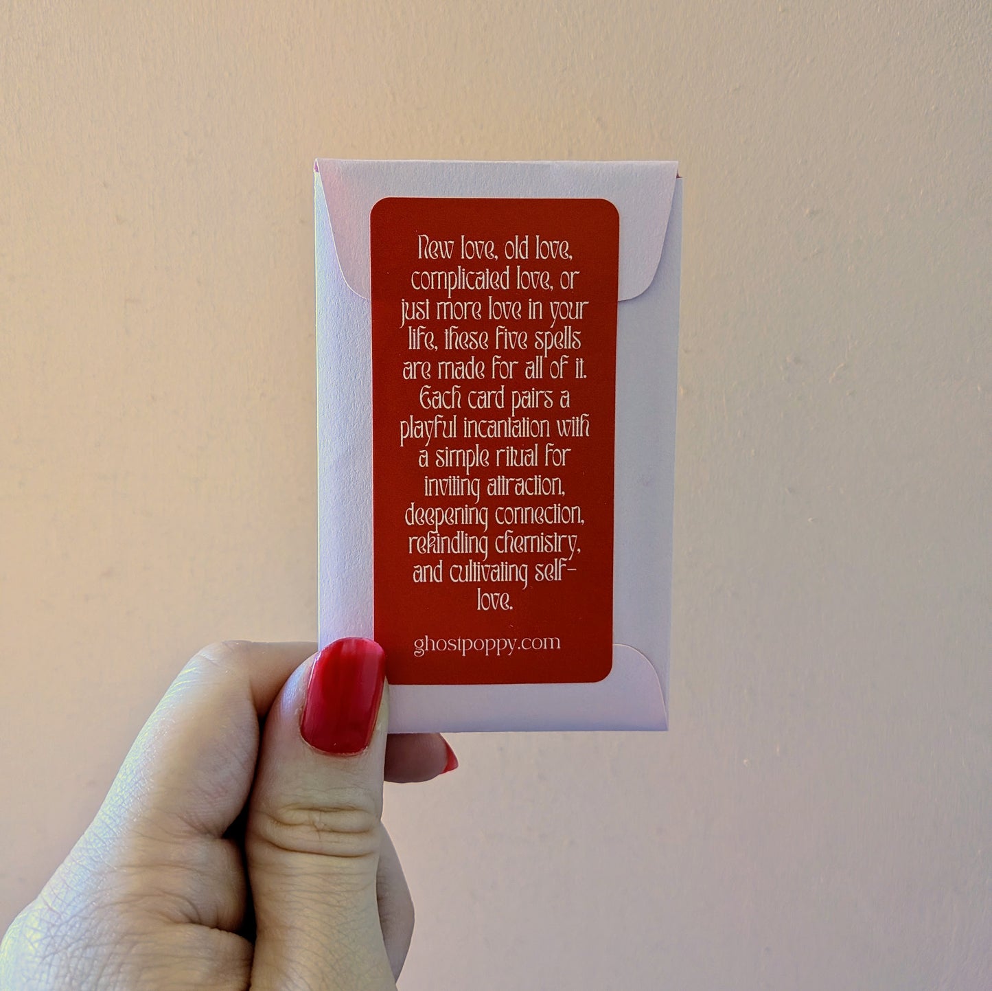 A hand holds a small envelope with a red label for a deck of Love Spell Cards featuring playful incantations and simple rituals to invite more love into your life.