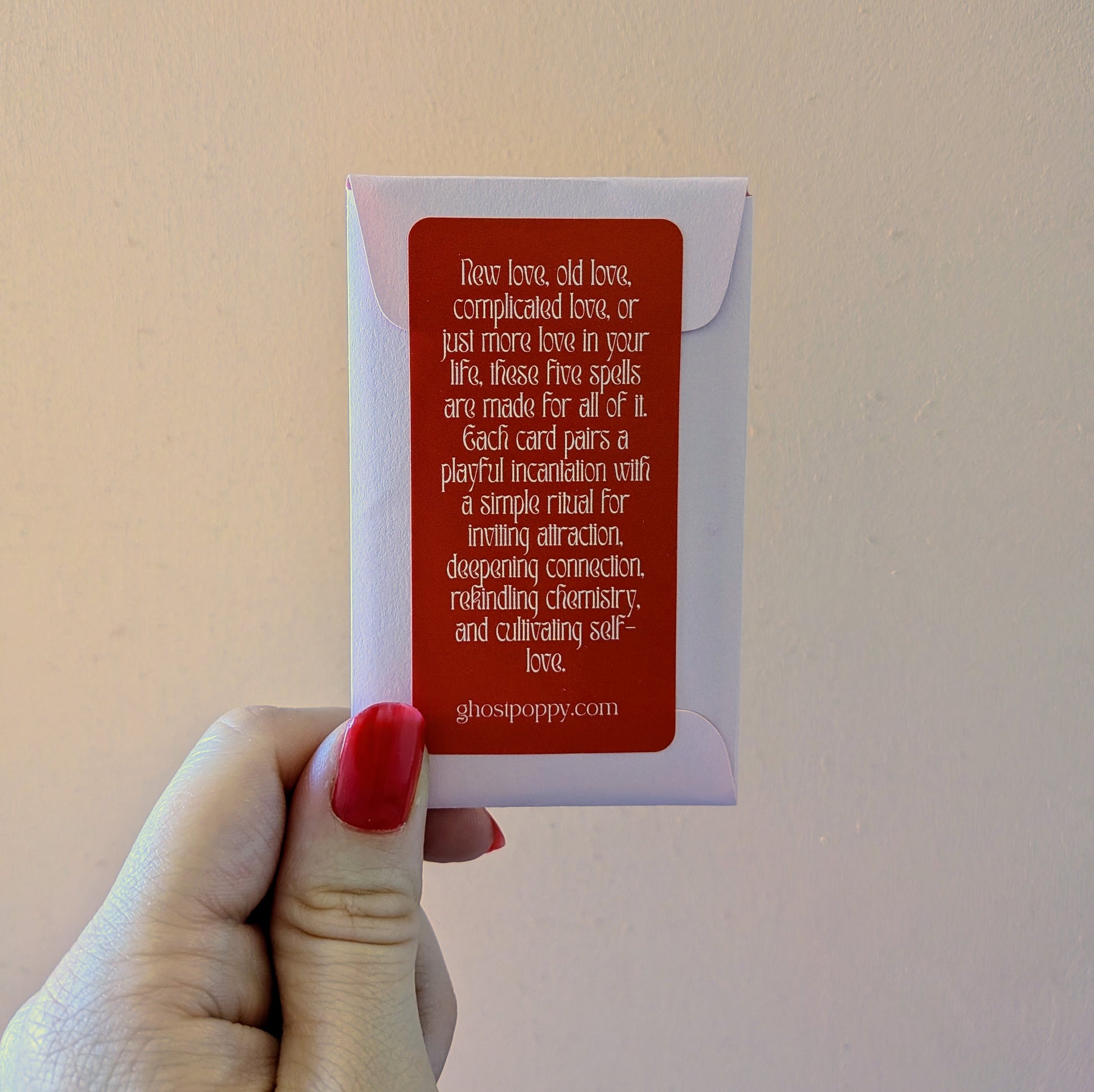 A hand holds a small envelope with a red label for a deck of Love Spell Cards featuring playful incantations and simple rituals to invite more love into your life.