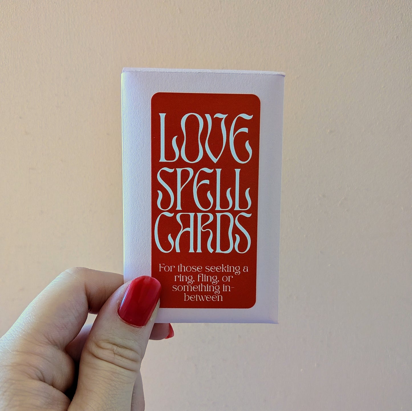 A hand holds a deck of Ghost Poppy Love Spell Cards with a red label that reads, For those seeking a ring, fling, or something in-between.