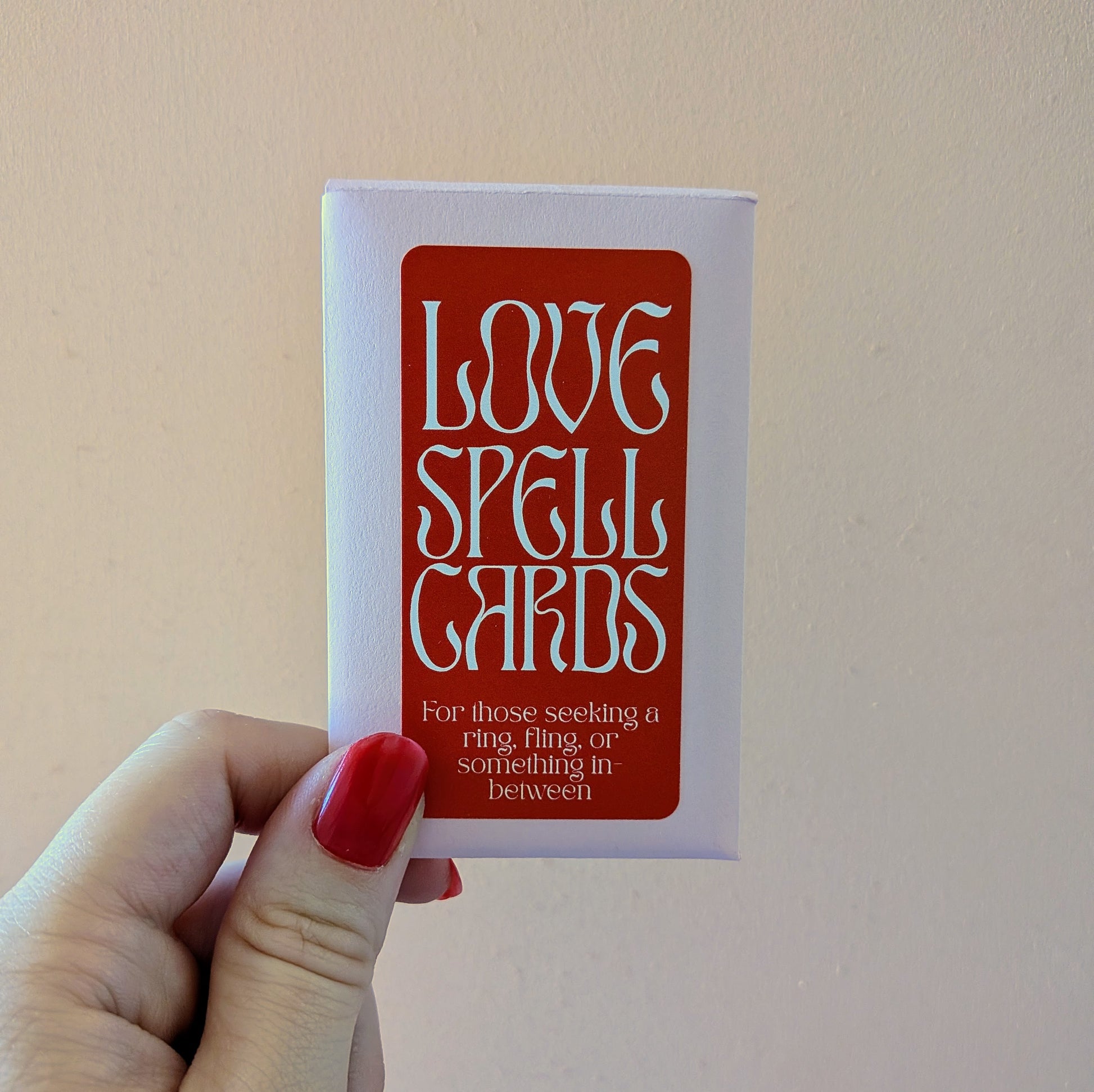 A hand holds a deck of Ghost Poppy Love Spell Cards with a red label that reads, For those seeking a ring, fling, or something in-between.