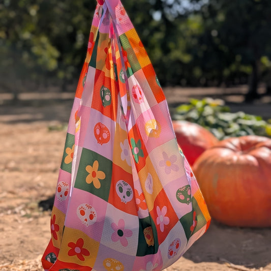 A Ghost Poppy Bloom & Burn Reusable Tote Bag featuring a checkerboard grid pattern of colorful skulls and flowers, hanging in an orchard with pumpkins in the background.