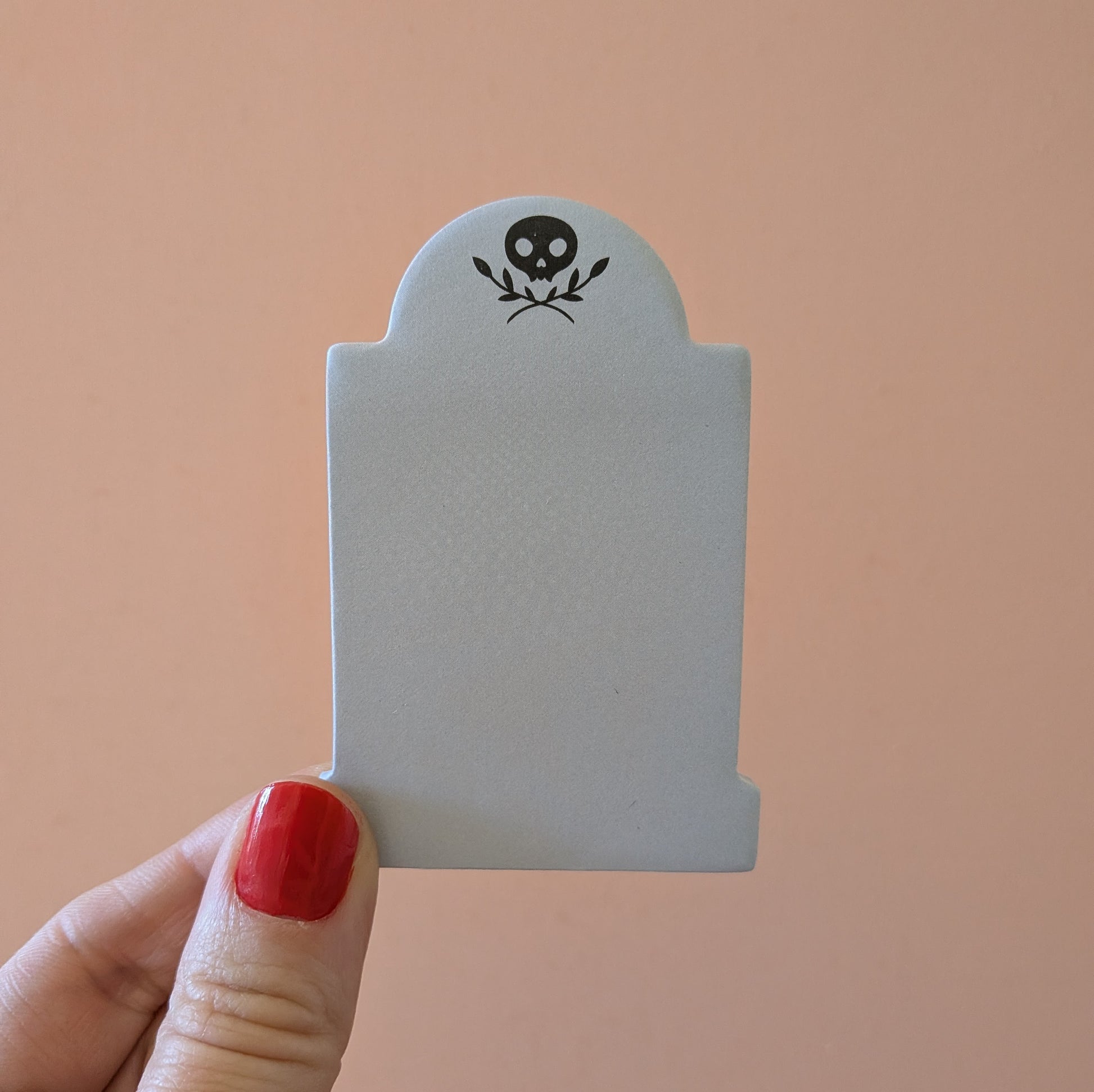 A hand holds a 3x2 inch tombstone-shaped Mini Memento Mori Sticky Note by Ghost Poppy, featuring a black skull and leaf icon at the top.