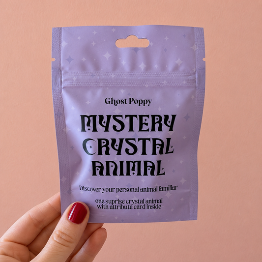 A hand holds a Ghost Poppy Mystery Crystal Animal packet against a peach background. The packet contains a surprise crystal animal figurine and an attribute card inside.