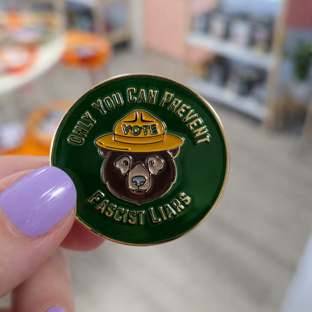Pin with bear design and text held by a hand in a blurred indoor setting