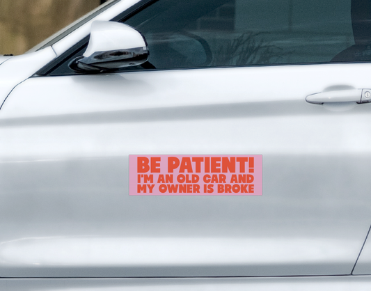 A pink rectangular Be Patient! Car Magnet on a silver car door that reads, BE PATIENT! IM AN OLD CAR AND MY OWNER IS BROKE. This weatherproof magnet is designed for long-lasting use.